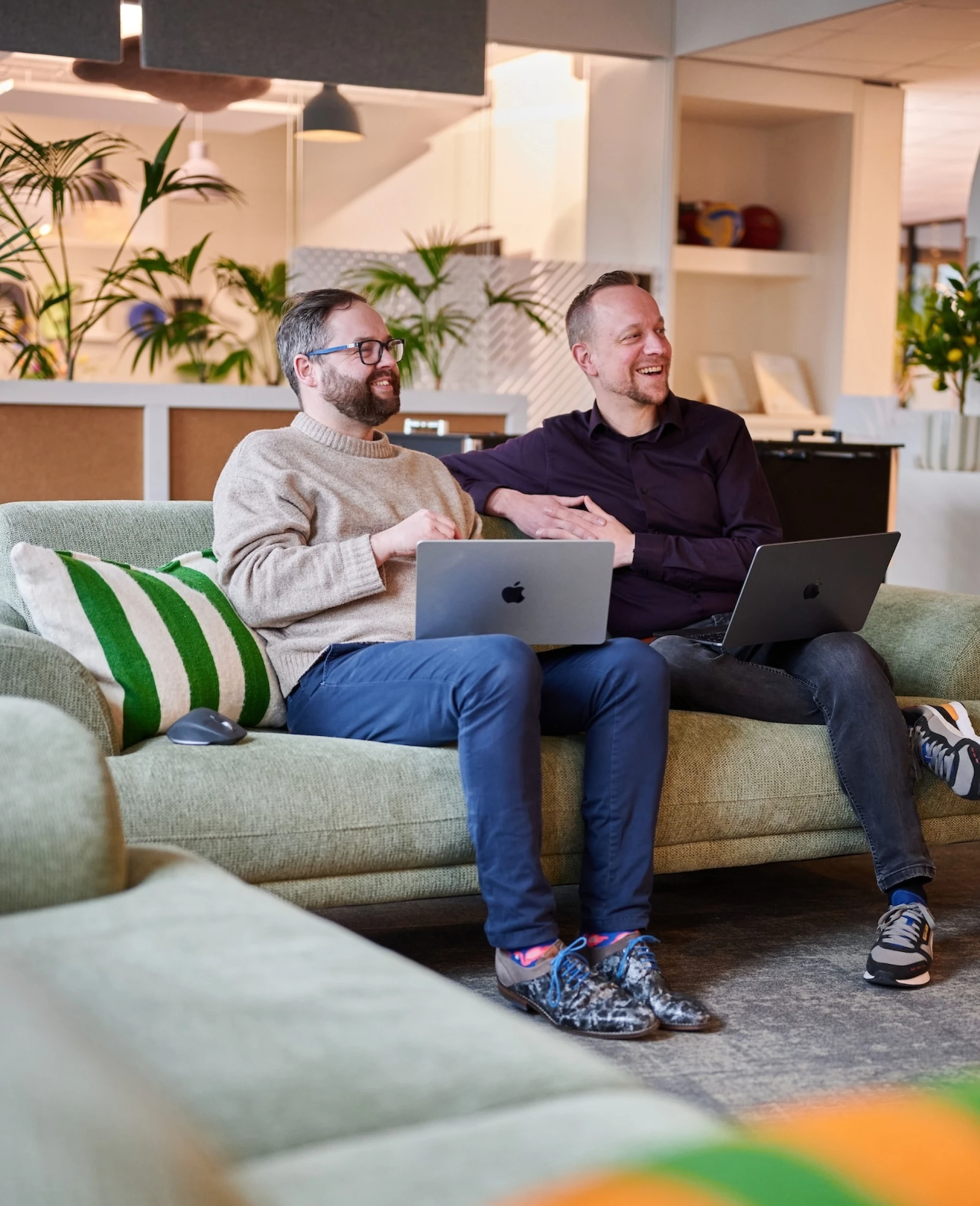 Twee mensen zitten op een bank met laptops in een gezellige ruimte.