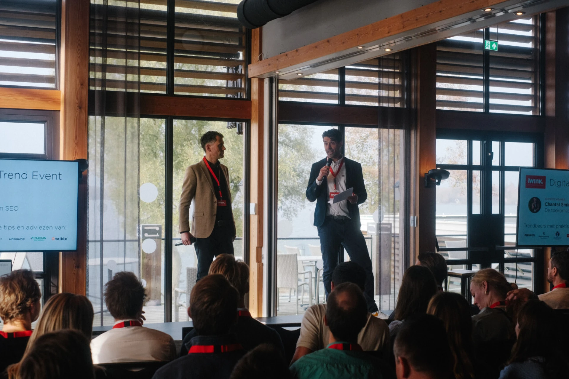 Twee sprekers presenteren op een podium tijdens een conferentie.