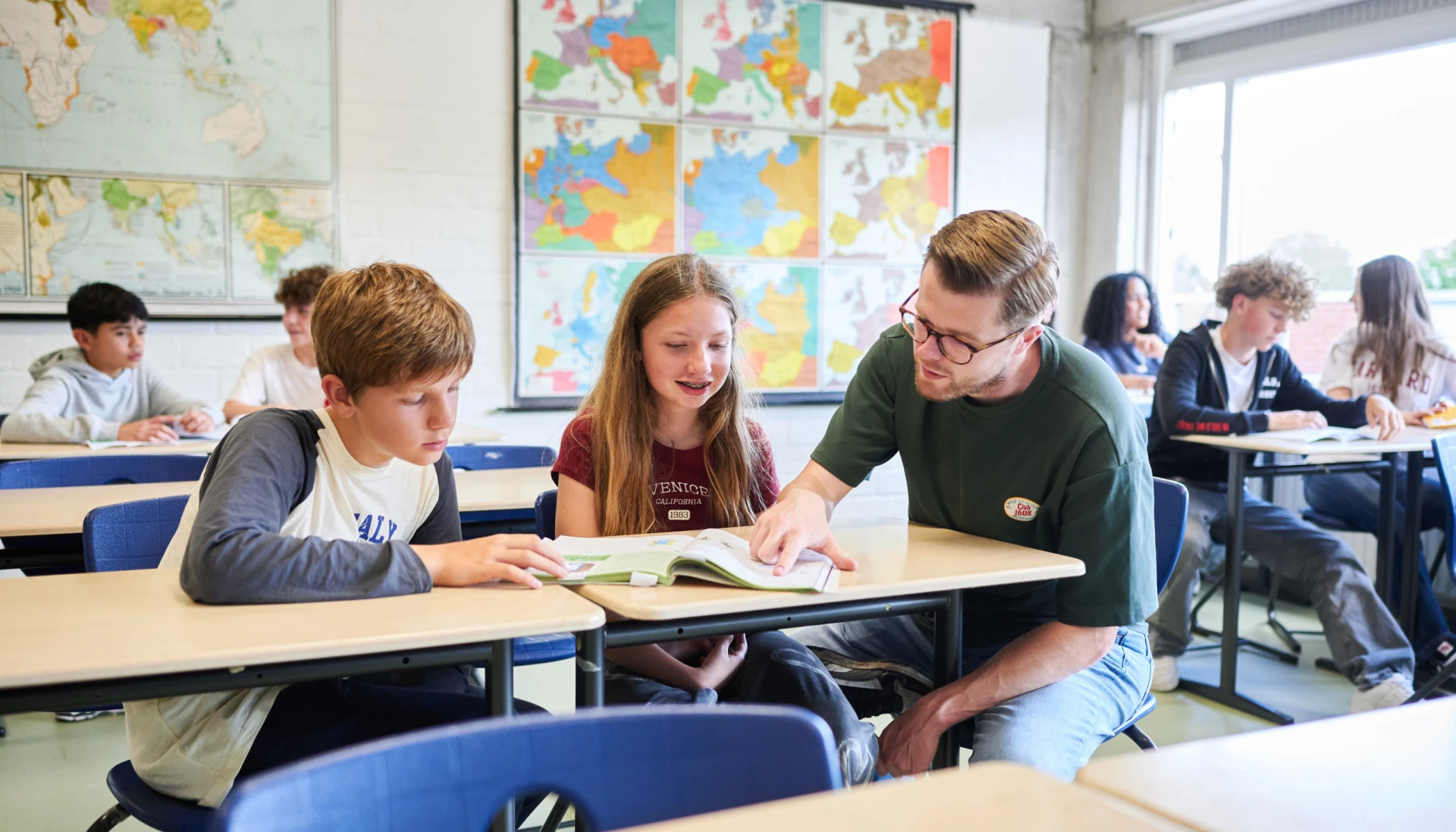 Een leraar helpt twee leerlingen bij het lezen van een boek in de klas.