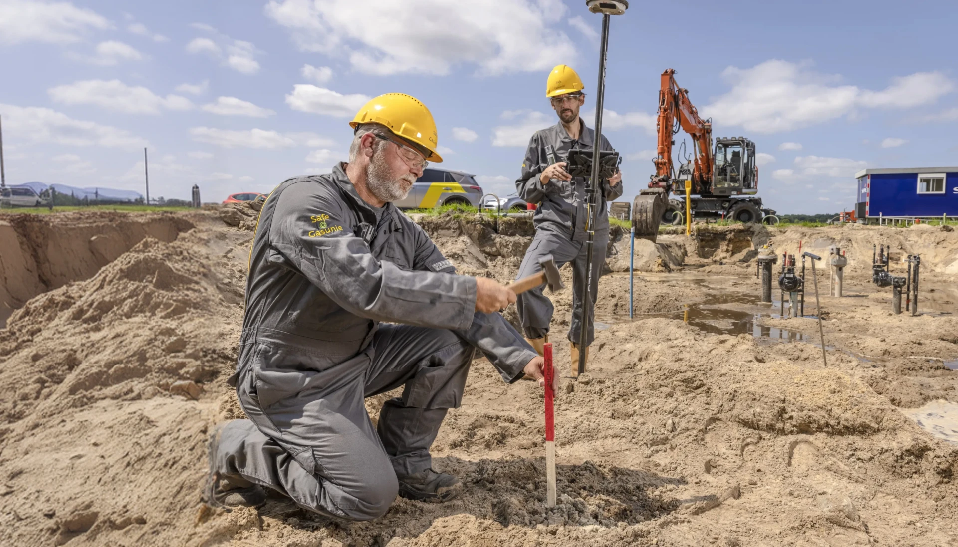 Twee bouwvakkers aan het werk op een bouwplaats met graafmachine op de achtergrond.