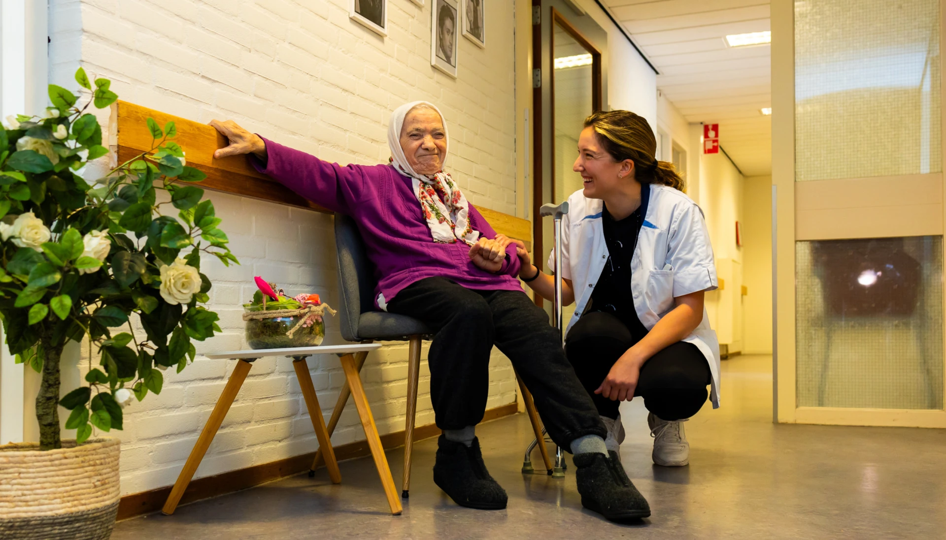 Een oudere vrouw zit op een bank terwijl een verzorgster glimlachend naast haar knielt in een gang.