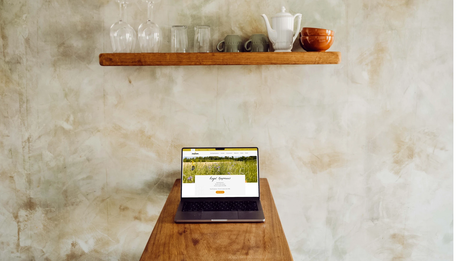 Een laptop op een houten tafel met een plank erboven met glazen en een theepot.