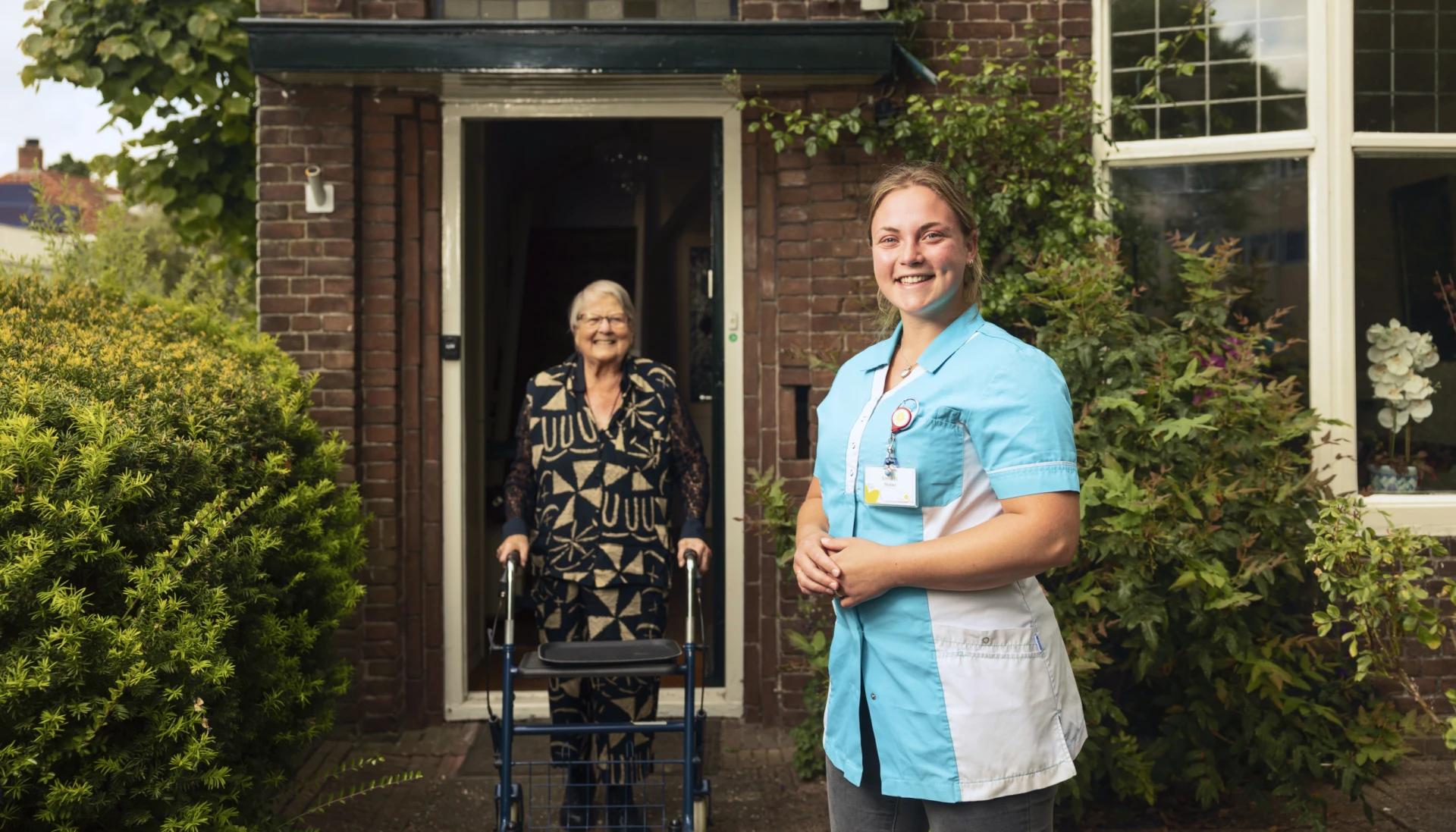Verzorgende helpt oudere vrouw met rollator voor een huis.