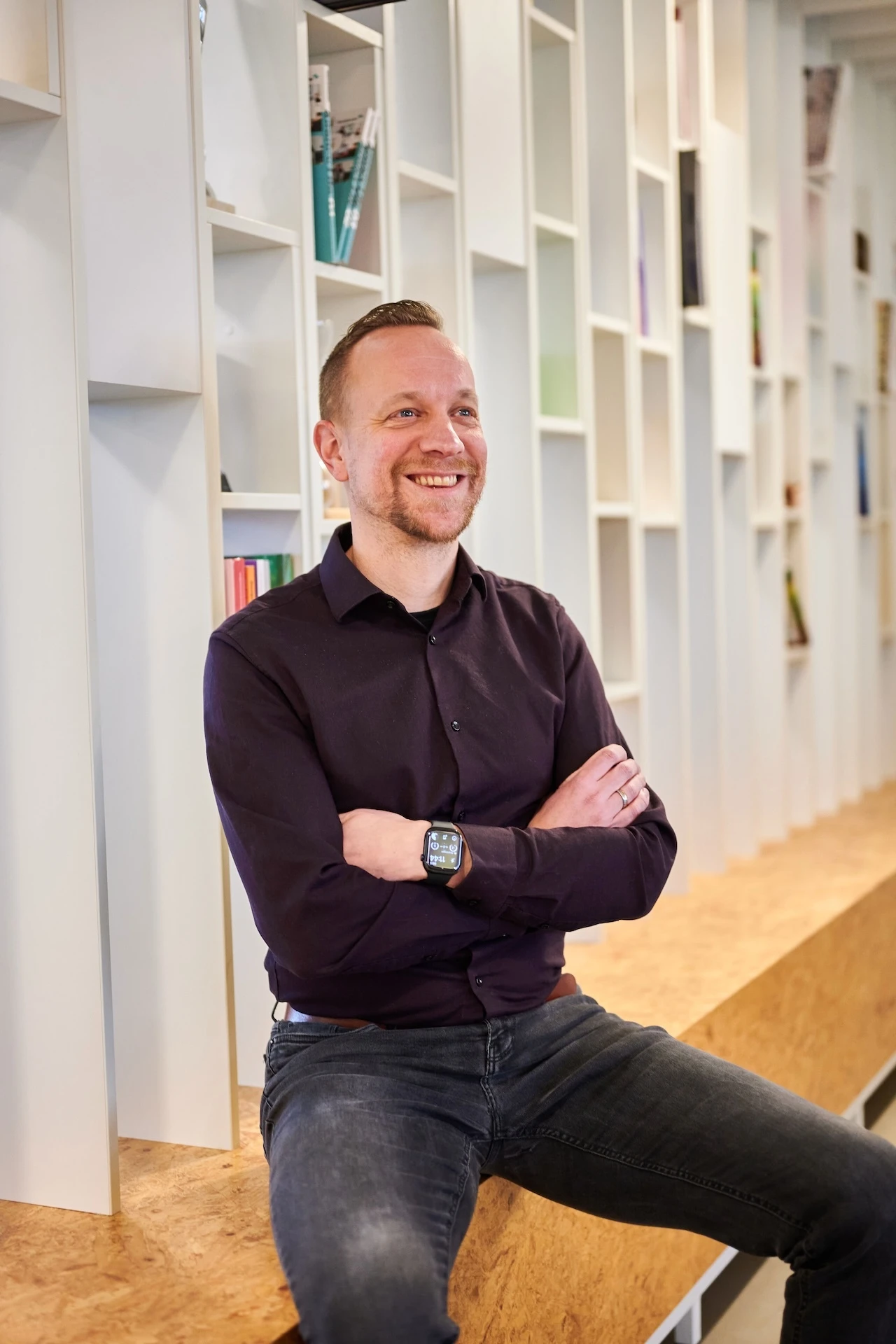 Een lachende man zit met gekruiste armen voor een moderne boekenkast.