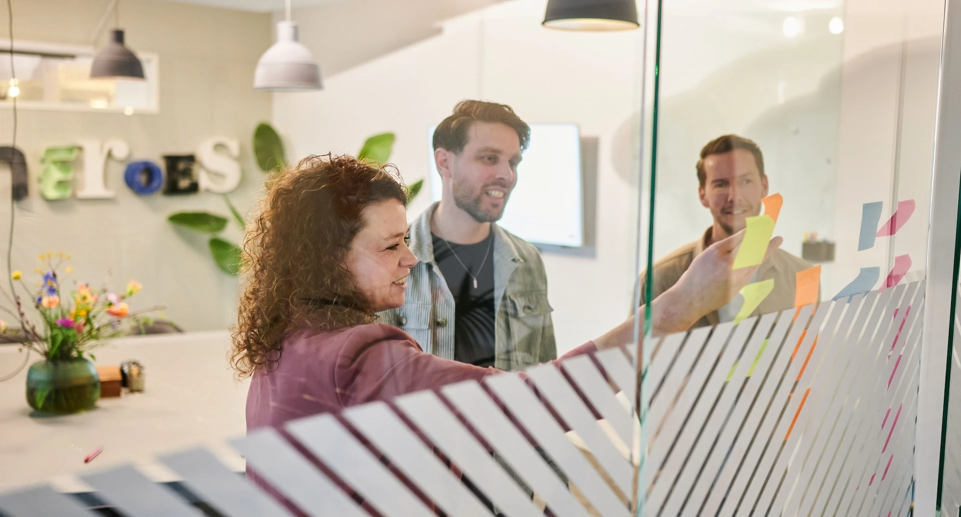 Mensen werken samen aan een glazen muur met plaknotities in een kantooromgeving.