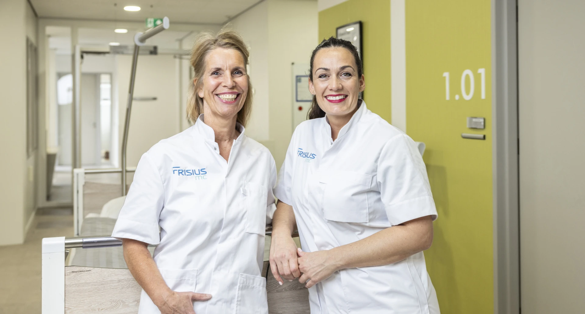 Twee vrouwelijke medische professionals in witte uniformen lachend in een gang.