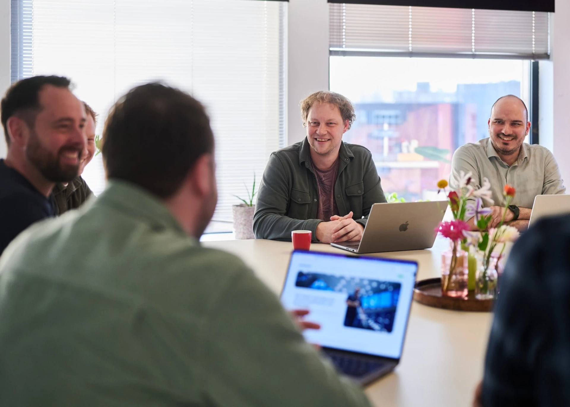 Mensen in vergadering rond een tafel met laptops en bloemen.