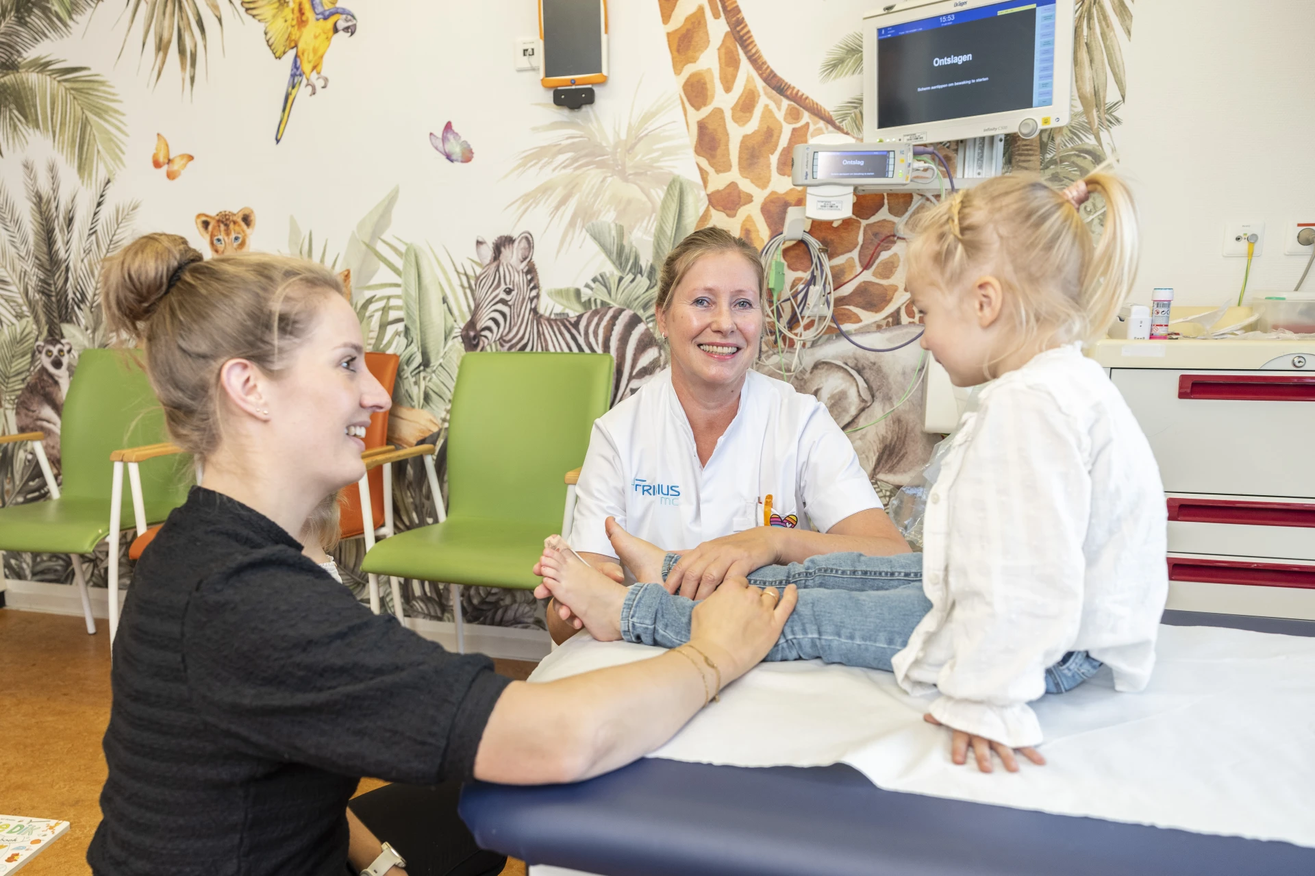 Een verpleegkundige praat met een kind en een ouder in een kleurrijk ingerichte onderzoekskamer.