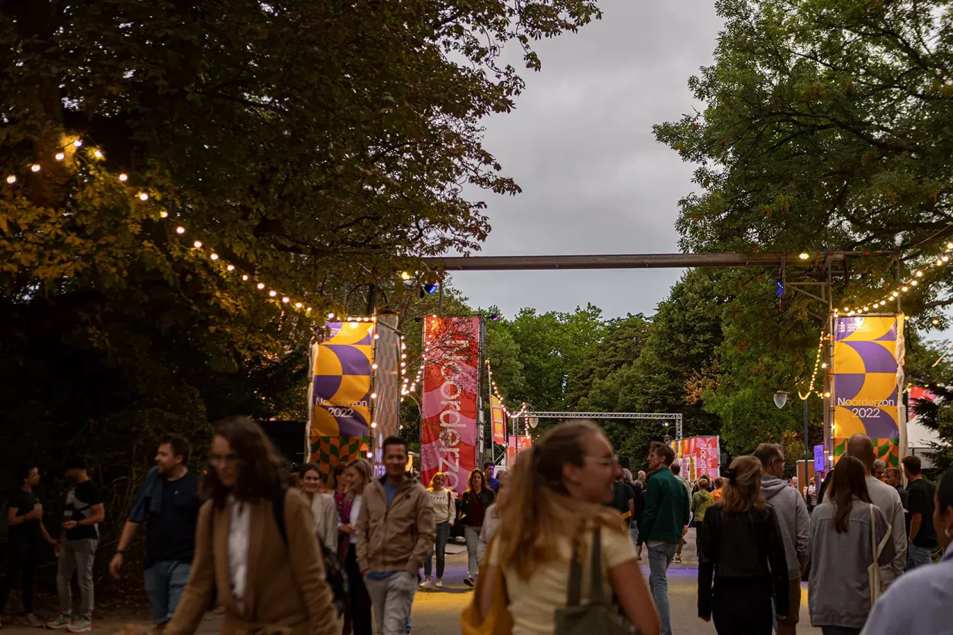 Een drukbezochte festivalingang met decoratieve bogen en sfeerverlichting omringd door bomen.