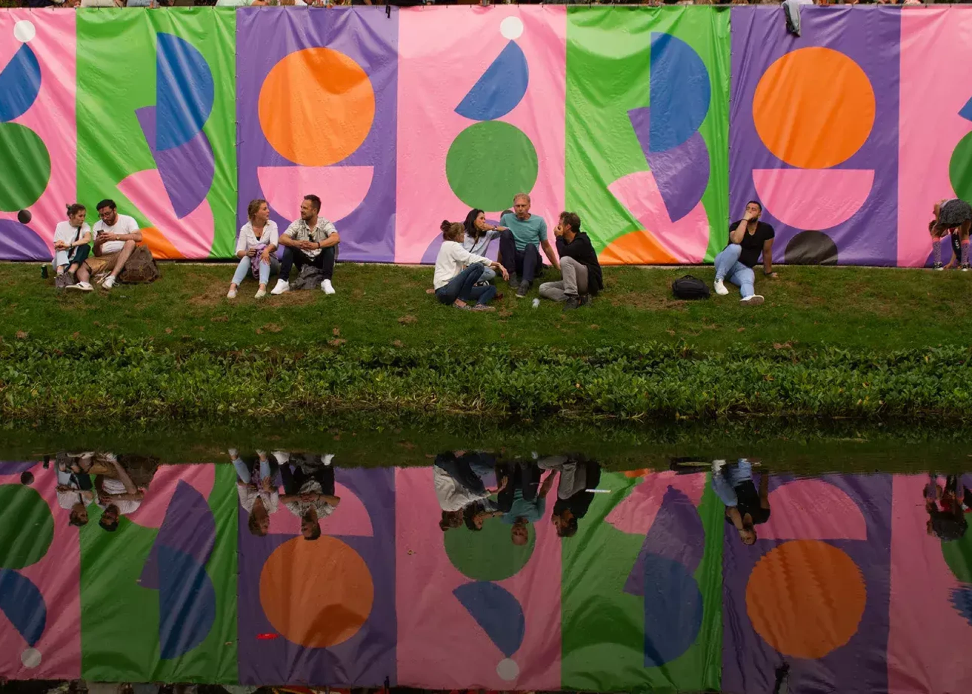 Mensen zitten bij een kleurrijk kunstwerk langs het water.