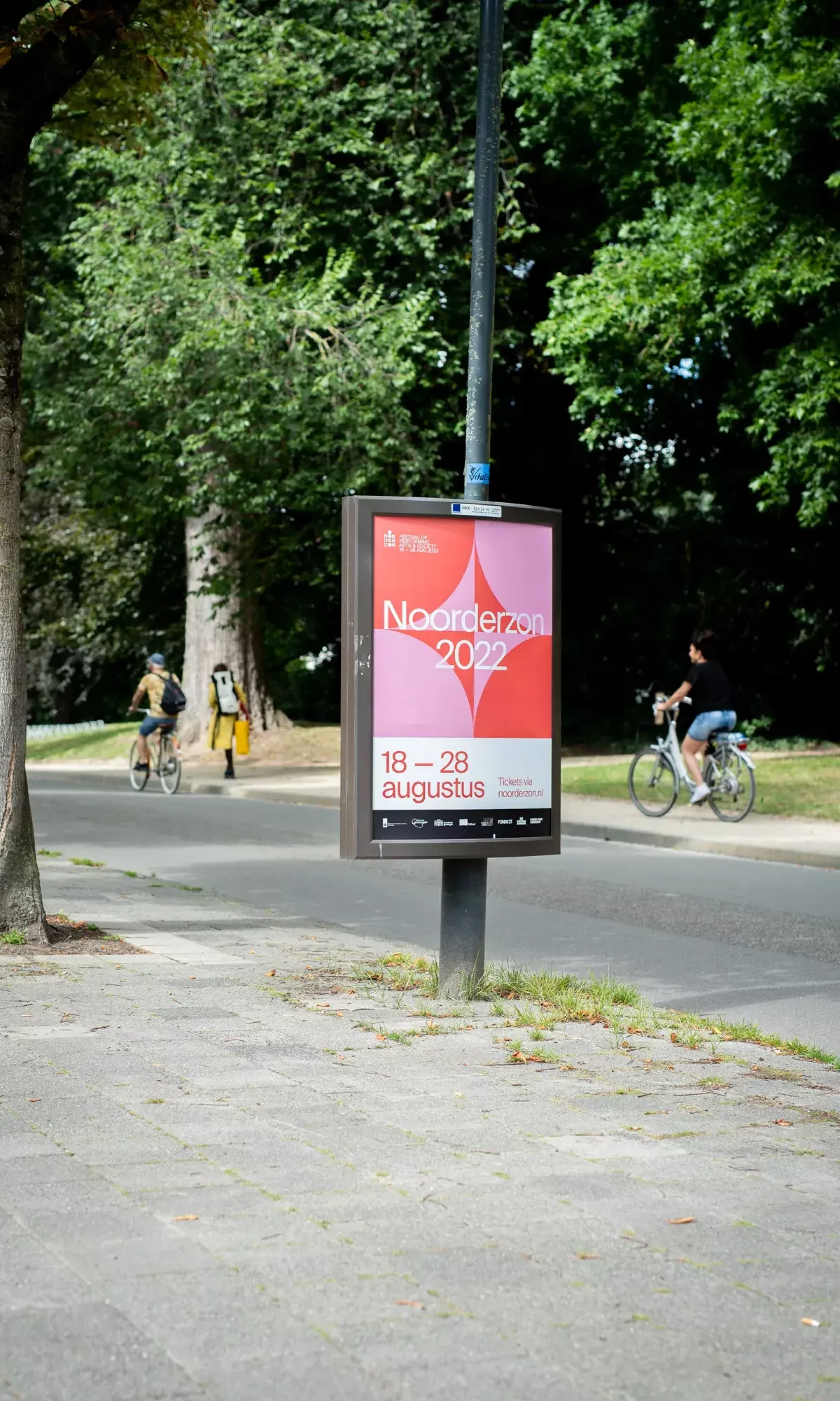 Fietsers op een pad naast een bord met een evenementposter in een groene omgeving.