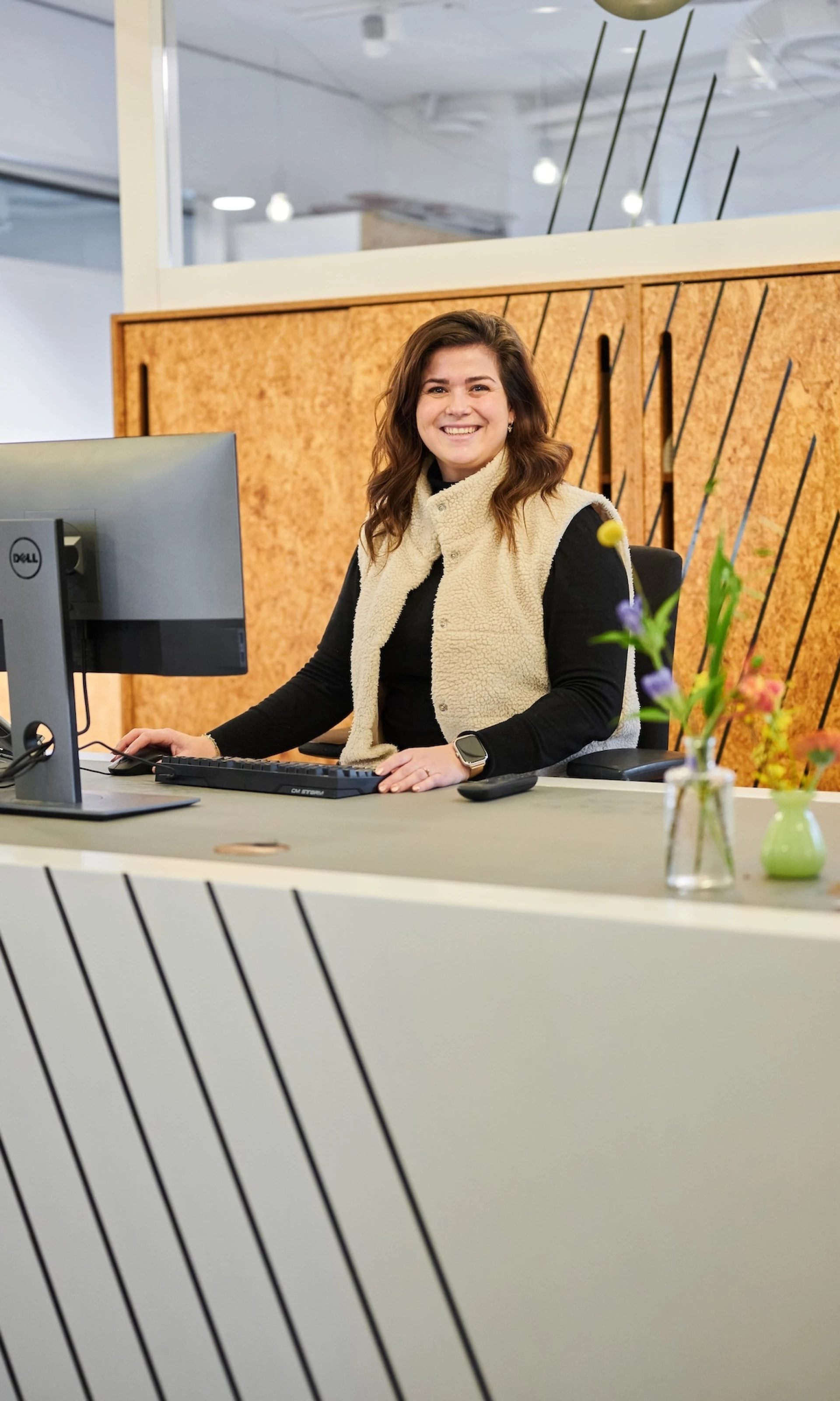 Vrouw bij een bureau met computer, glimlachend naar de camera.