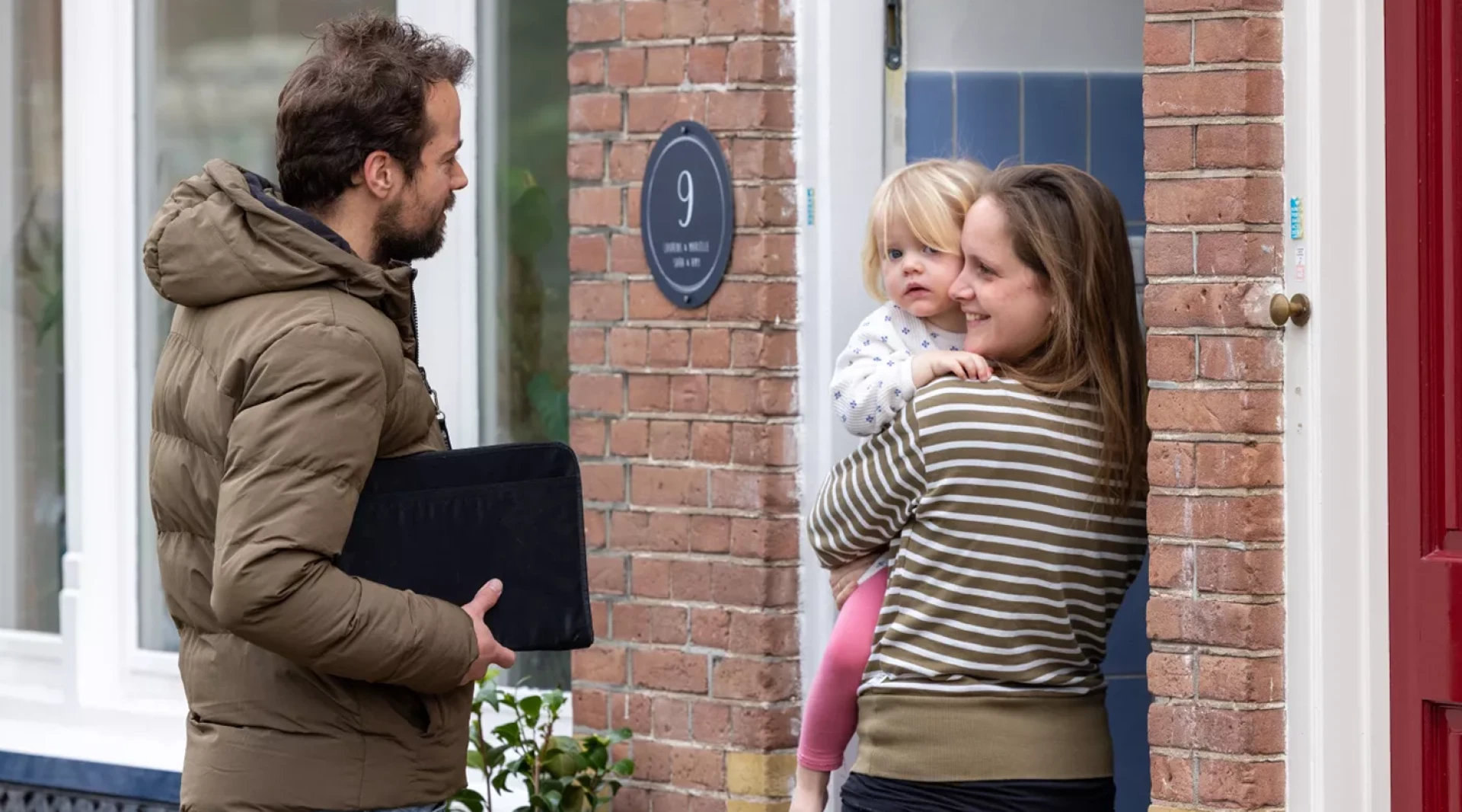 Een man ontmoet een vrouw die een kind vasthoudt bij de voordeur van een huis.