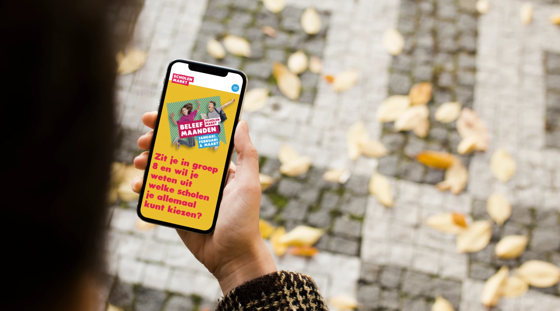 Persoon bekijkt telefoon met kleurrijke afbeelding en tekst over stemmen en diversiteit, tijdens een wandeling op een met bladeren bedekte stoep.