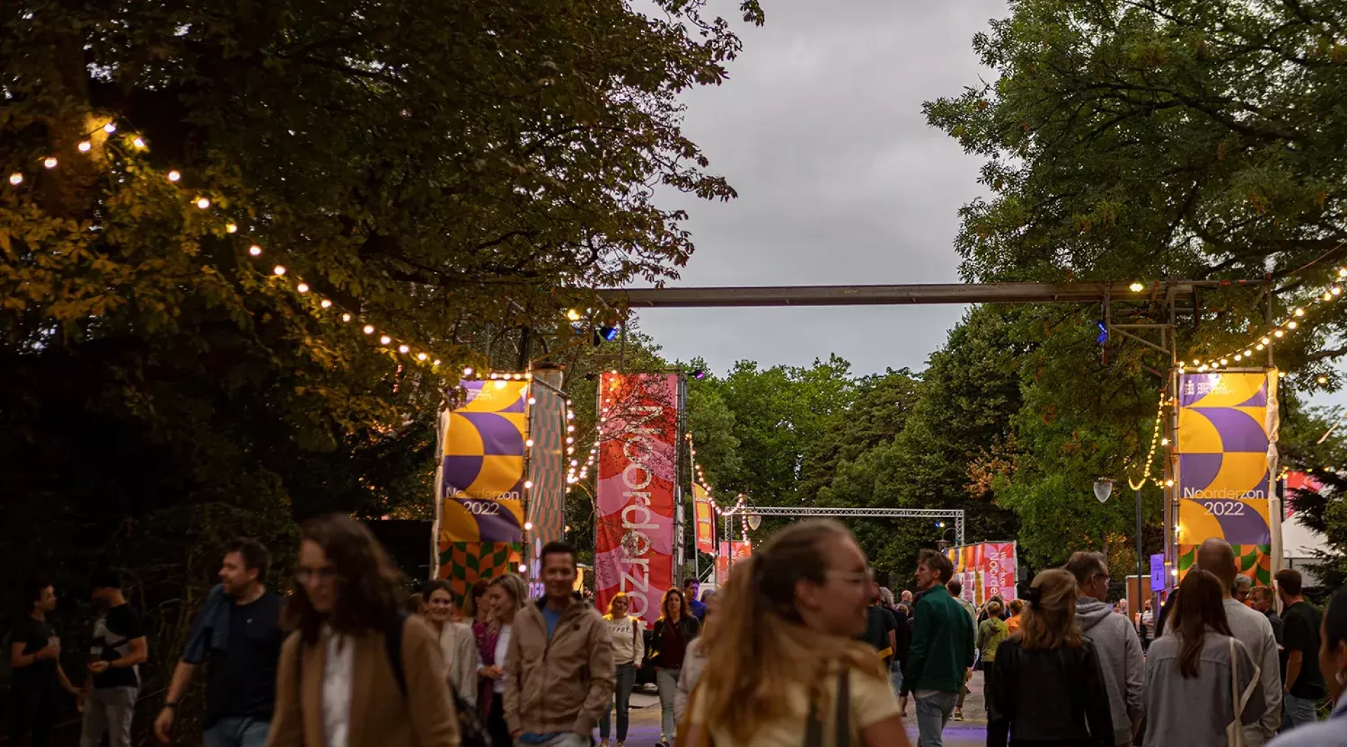 Een drukbezochte festivalingang met decoratieve bogen en sfeerverlichting omringd door bomen.