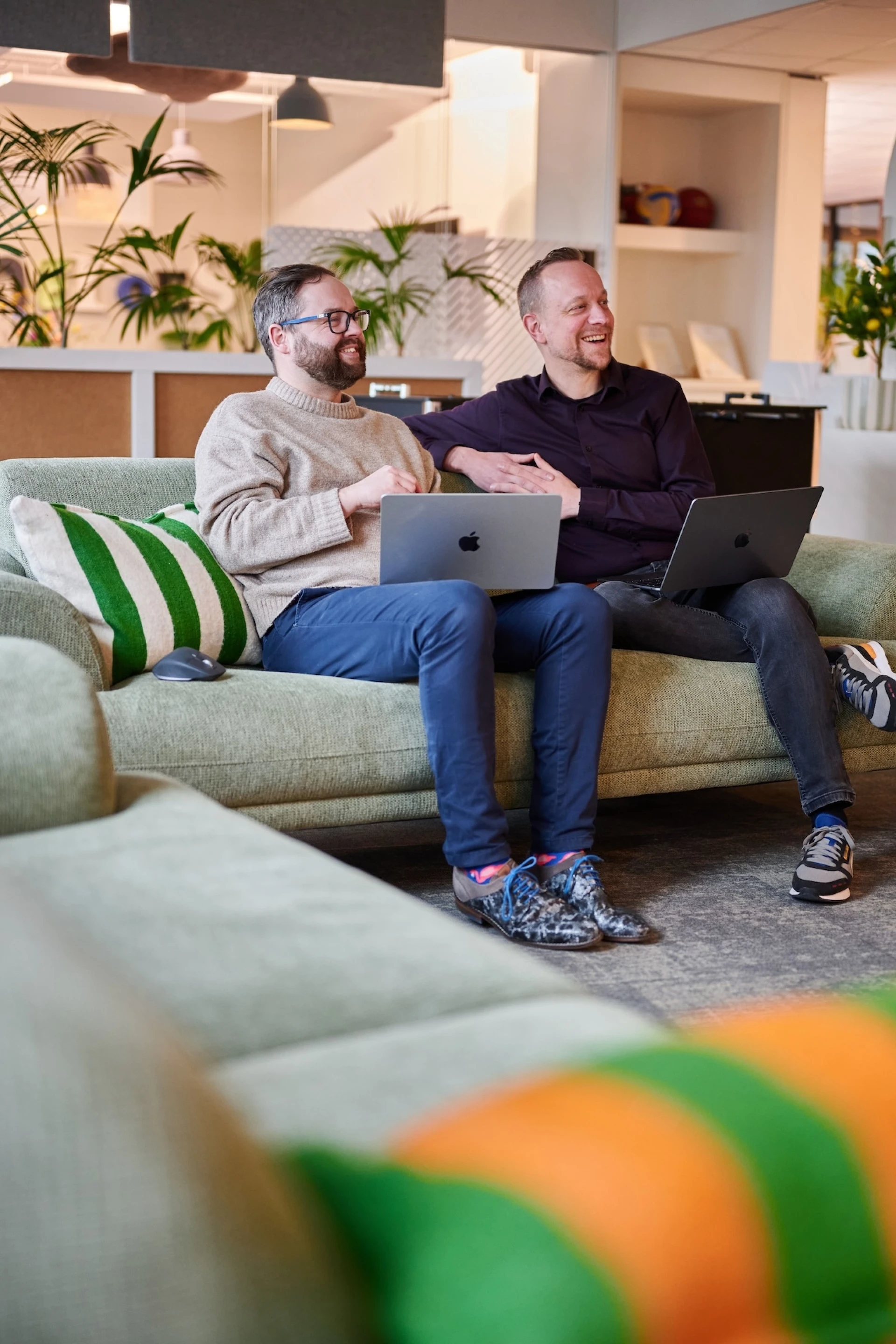Twee mensen zitten op een bank met laptops in een gezellige ruimte.