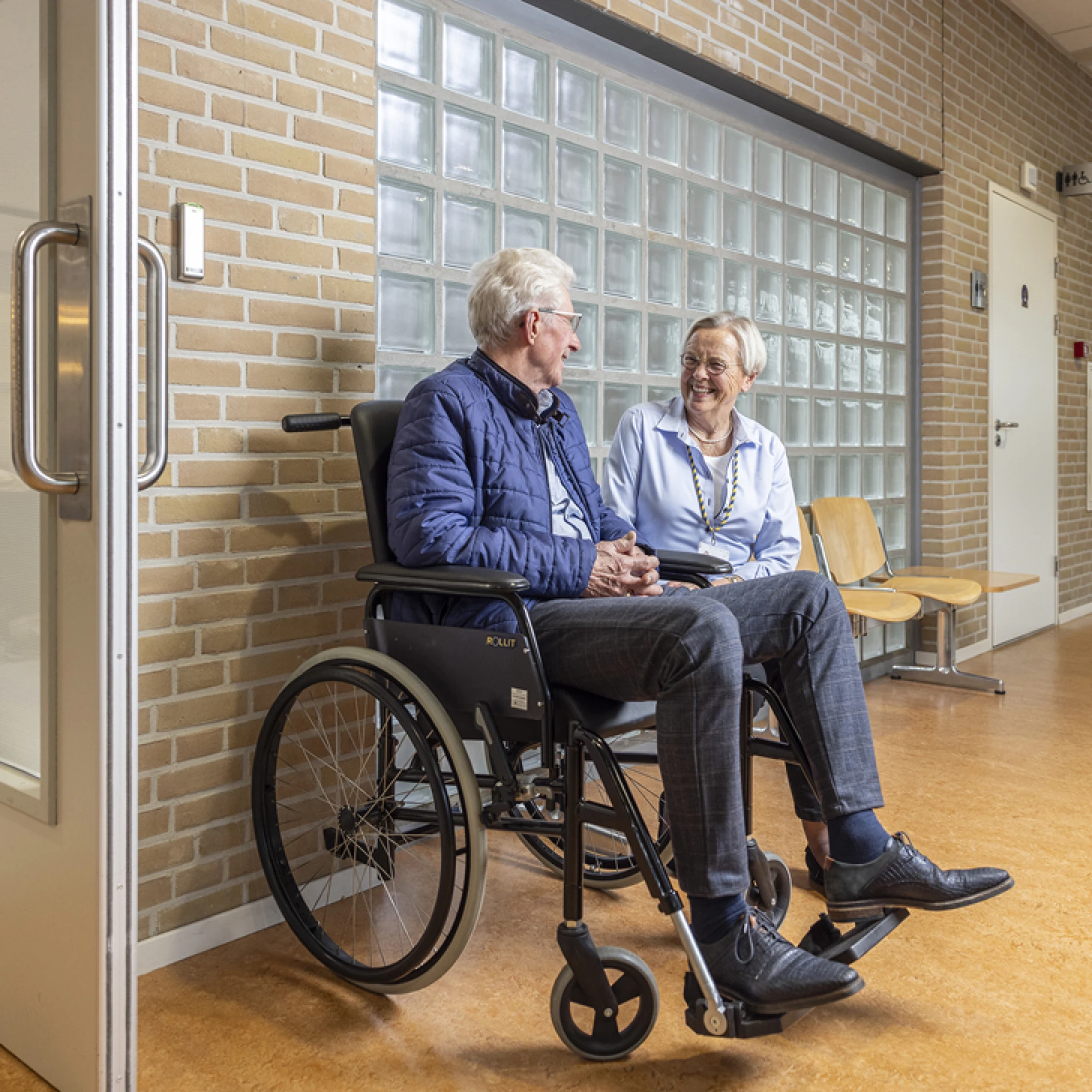 Twee mensen in gesprek in een gang, één persoon zit in een rolstoel.